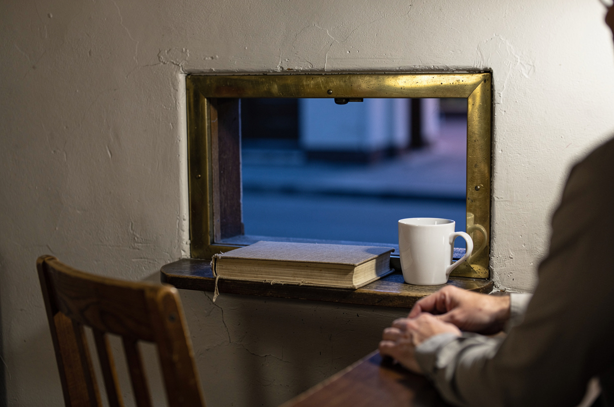 Of the inside of a small early-1900s bank teller window at dawn: a plain unmarked brass-framed opening in a plaster wall, a single closed ledger resting on the wooden ledge, a plain unmarked ceramic mug beside it, and an empty chair turned slightly away. Cool morning light from the street beyond, real texture in the plaster, the brass and the worn wood.