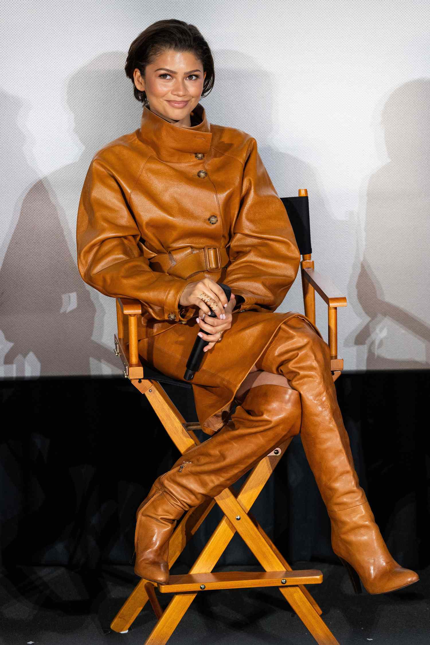 Zendaya seated in a director's chair in a tan leather outfit, holding a microphone, on set.